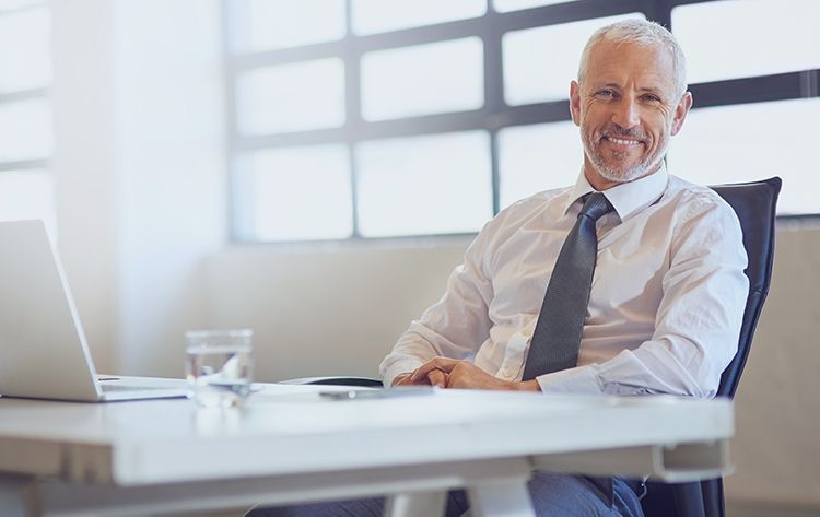 Lächelnder Geschäftsführer sitzt entspannt im Büro Älterer Mann im Anzug sitzt lächelnd an einem Schreibtisch im modernen Büro – symbolisiert Erfahrung, Unternehmensleitung und Führungsrolle